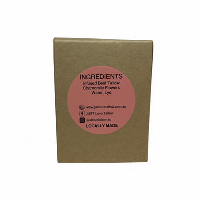 A square brown kraft paper box standing upright against a light gray fabric backdrop, featuring a circular pink label on the front with black text listing ingredients and the brand JUST Love Tallow.
