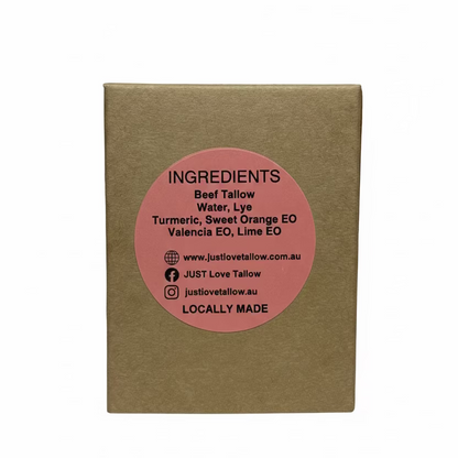 A square brown kraft paper box with a circular pink label on the front, photographed against a light gray fabric backdrop. The label reads 'INGREDIENTS' and lists natural tallow, lye, turmeric, sweet orange EO, and lima EO, with the brand name justlovetailow visible.